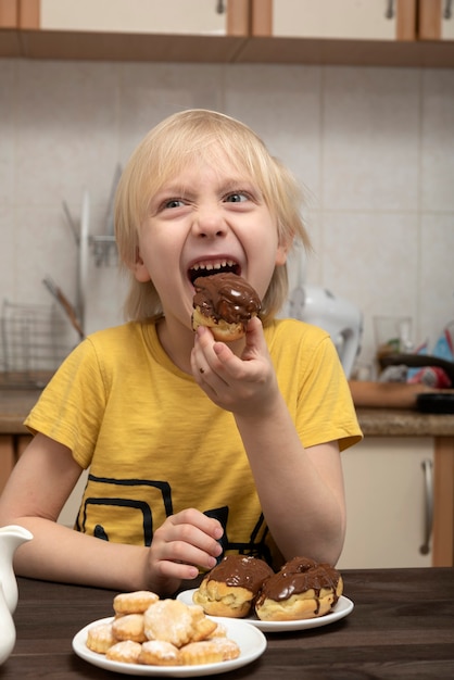 Szczęśliwy blond chłopiec je ciasto. Dziecko zjada ekler. Rama pionowa.