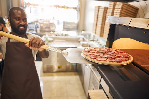 Szczęśliwy brodaty mężczyzna w fartuchu robi pizzę i wkłada ją do piekarnika do pieczenia for