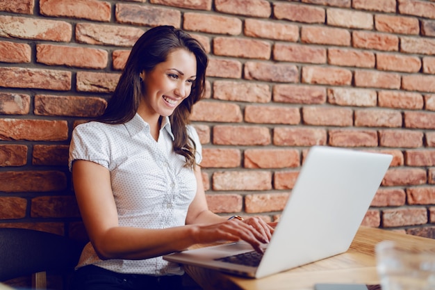 Szczęśliwy całkiem kaukaski brunetka siedzi w kawiarni i za pomocą laptopa. Ręce są na klawiaturze. W tle jest mur z cegły.