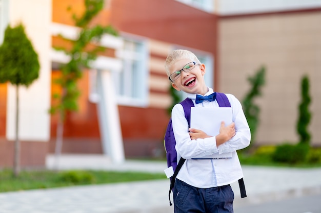 Szczęśliwy chłopiec blond uczeń w okularach z plecakiem i białą książką stoi w szkole i się śmieje