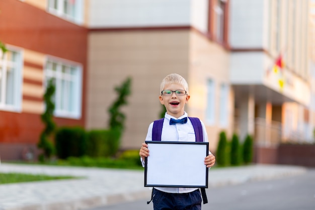 Szczęśliwy chłopiec uczeń w blond okularach i plecaku stoi przy szkole i trzyma znak z białym prześcieradłem.