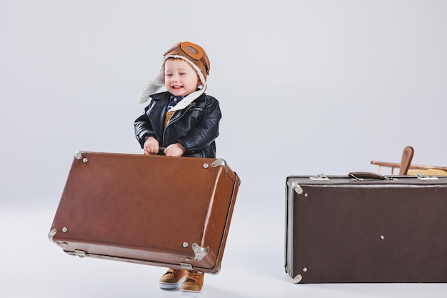 Szczęśliwy chłopiec w kasku i kurtce pilota niesie brązową walizkę Portret dziecka pilot dziecko w skórzanej kurtce Mały podróżnik z walizką