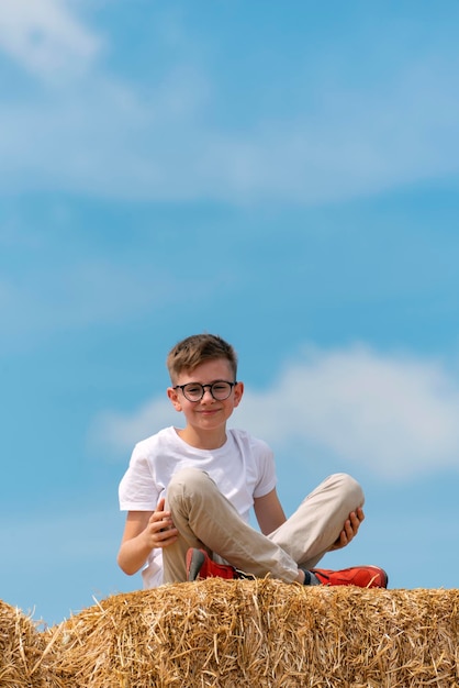 Szczęśliwy chłopiec w okularach siedzący na beli siana na tle błękitnego nieba latem Rama pionowa Beztroskie dzieciństwo