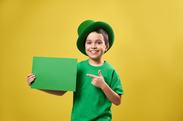 Szczęśliwy chłopiec w zielonych ubraniach i irlandzkim kapeluszu przykłada palec wskazujący do kartki papieru z miejscem na kopię i uśmiecha się, pozując na żółtej powierzchni