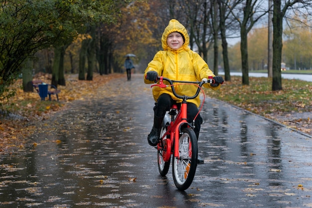 Szczęśliwy chłopiec w żółtym płaszczu jeździ na rowerze po mokrej alejce w deszczu. Jesienny dzień