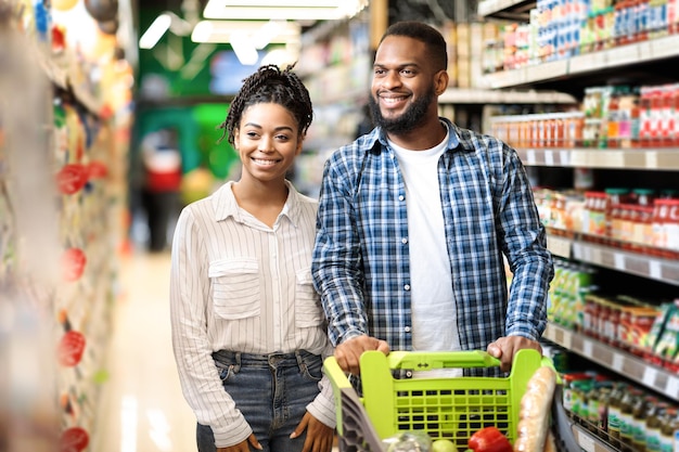 Szczęśliwy czarny mężczyzna i kobieta w supermarkecie, afroamerykańska rodzina robi zakupy razem na zakupy spożywcze kupowanie jedzenia spacery, pchanie wózka wzdłuż półek sklepowych. Konsumpcjonizm, reklama w supermarkecie