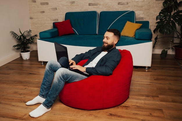 Szczęśliwy człowiek freelancer pracuje na laptopie siedząc na czerwonym fotelu w domu.