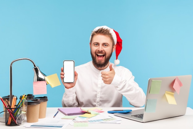 Szczęśliwy człowiek pozytywny w Santa Claus kapelusz i białą koszulę Wyświetlono kciuk w górę gest trzymając pusty wyświetlacz smartphone siedząc w miejscu pracy, zakupy online. Kryty studio strzał na białym tle na niebieskim tle