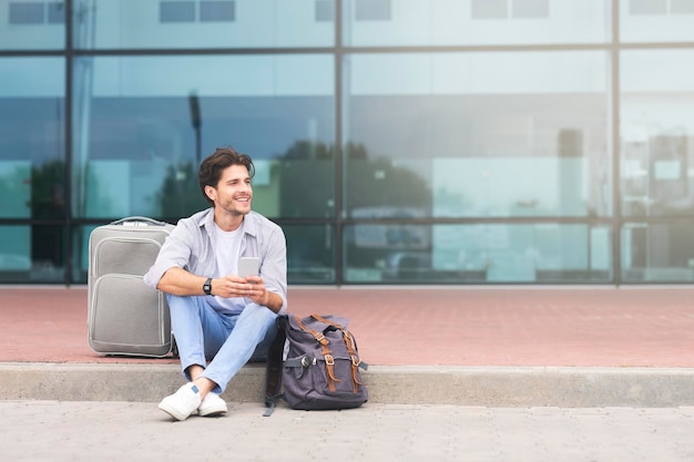 Szczęśliwy człowiek siedzący z bagażem i smartfonem w pobliżu terminalu lotniska