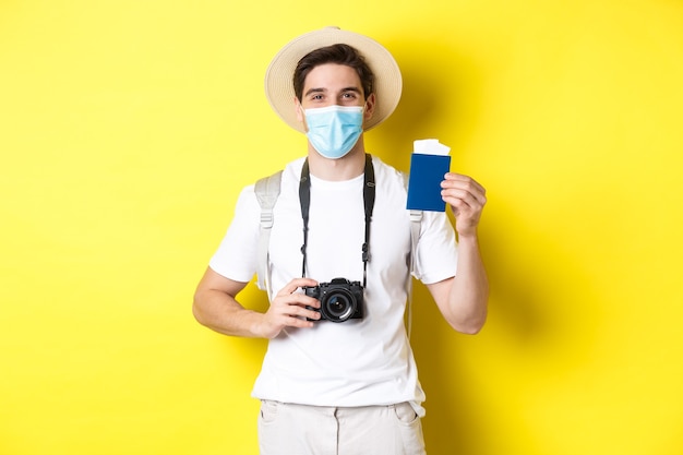 Szczęśliwy człowiek turysta z przodu, pokazując paszport i bilety na wakacje, udaje się w podróż podczas pandemii, żółta ściana