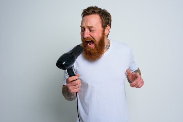 Szczęśliwy człowiek z brodą używa suszarki do włosów jako mikrofonu i tańczy