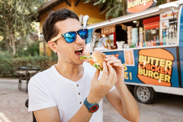 Szczęśliwy człowiek zjada hamburgera w pobliżu foodtrucka na świeżym powietrzu. Koncepcja Streetfood i fast foodów