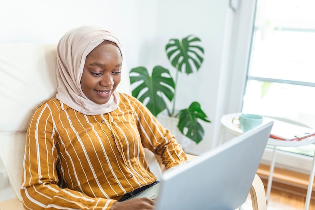 Szczęśliwy Dość Muzułmańskiej Afryki Za Pomocą Laptopa Siedząc Na Wygodnej Kanapie. Piękna Młoda Muzułmanka Korzysta Z Laptopa I Uśmiecha Się Siedząc Na Kanapie W Domu