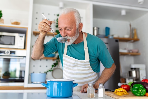 Szczęśliwy emerytowany starszy mężczyzna gotujący w kuchni Koncepcja ludzi hobby emerytalnych Portret uśmiechniętego starszego mężczyzny trzymającego łyżkę, aby spróbować jedzenia