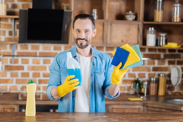 Szczęśliwy energiczny mężczyzna sprzątacz trzymając się za środki czyszczące, uśmiechając się i ubierając w rękawiczki