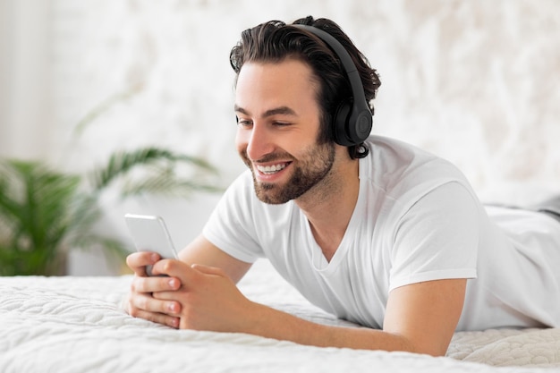 Szczęśliwy facet korzystający z wnętrza sypialni smartfona i zestawu słuchawkowego