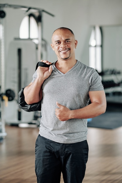 Szczęśliwy fit mieszanej rasy mężczyzna stojący w siłowni z torbą i pokazujący kciuki do góry