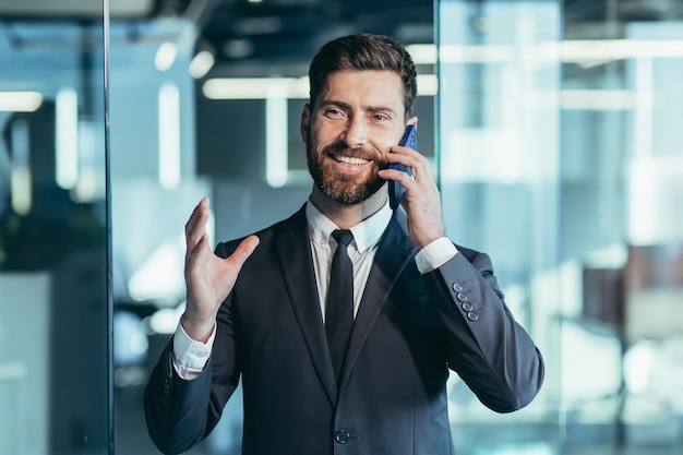 Szczęśliwy i uśmiechnięty biznesmen bawi się rozmawiając przez telefon, mężczyzna w nowoczesnym biurze, zbliżenie portretowe