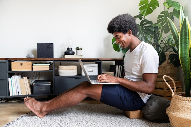 Szczęśliwy i uśmiechnięty młody człowiek african american pisania na laptopie w swoim przytulnym, ciepłym mieszkaniu. Skopiuj miejsce. Koncepcja domu. Technologia.