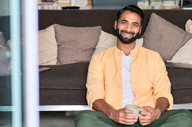 Zdjęcie szczęśliwy indyjski mężczyzna patrzący na kamerę pijącą kawę siedzącą na kanapie w domu