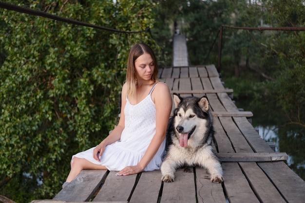 Szczęśliwy kaukaski blond kobieta w białej sukni pieścić alaskan malamute pies na moście wiszącym. miłość i przyjaźń między człowiekiem a zwierzęciem.