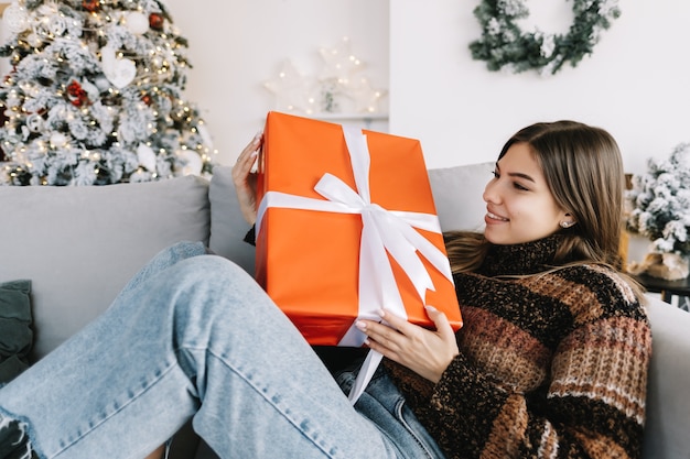 Szczęśliwy kaukaski kobieta trzyma duży prezent świąteczny siedząc na kanapie w domu w pobliżu choinki.