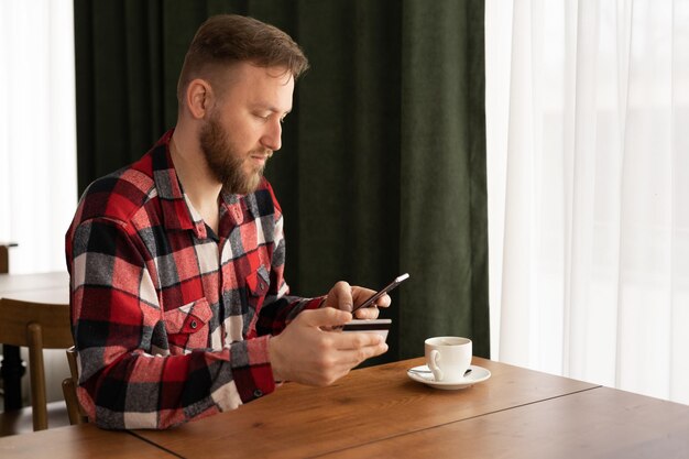 Zdjęcie szczęśliwy kaukazjski mężczyzna trzymający telefon komórkowy, wpisujący dane karty do dokonywania płatności online, siedzący w kawiarni, robiący zakupy online