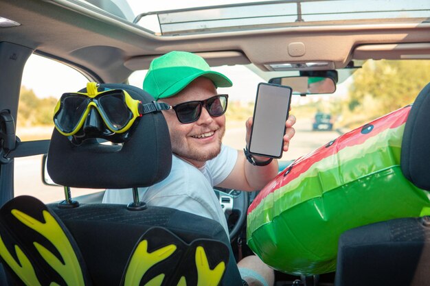 Szczęśliwy kierowca mężczyzna samochód pełen wakacyjnych rzeczy, trzymający telefon z białym ekranem