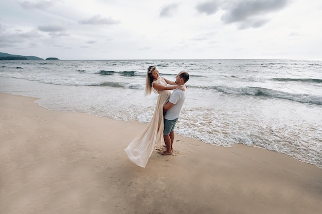 Szczęśliwy kochający mąż i żona ubrani na biało na egzotycznej plaży, mężczyzna przytula dziewczynę i podnosi ją z ziemi.