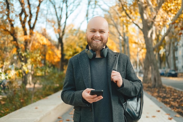 Szczęśliwy łysy brodaty mężczyzna spaceruje po ulicach jesienią, trzymając telefon