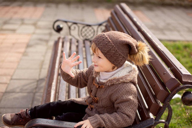 szczęśliwy mały blond chłopiec w brązowych ubraniach z dzianiny siedzi na ławce w parku