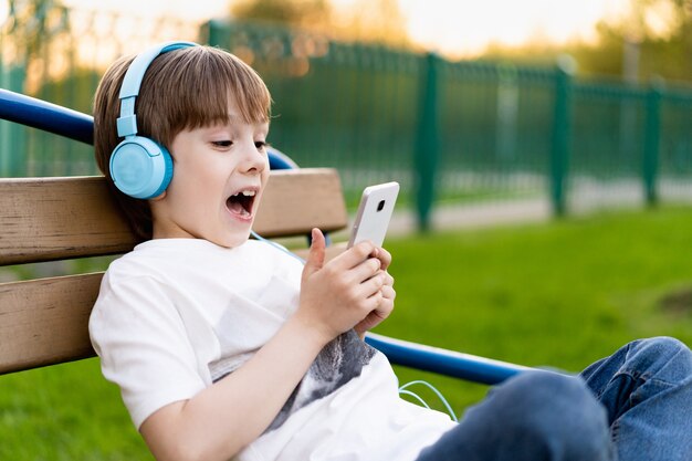 Szczęśliwy mały chłopiec na ulicy siedzi na ławce ze słuchawkami i telefonem i się uśmiecha