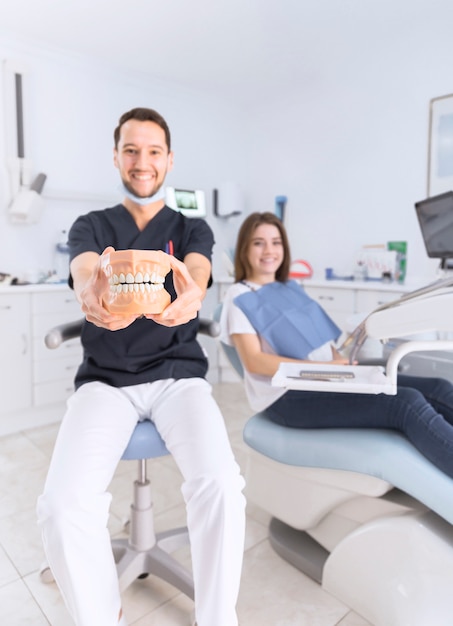 Zdjęcie szczęśliwy męski dentysta pokazuje zęby model siedzi przed żeńskim pacjentem przy stomatologiczną kliniką