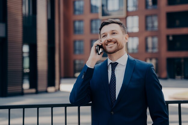 Szczęśliwy męski pracownik biurowy w niebieskim garniturze rozmawia przez telefon z kolegą lub partnerem o następnym projekcie biznesowym, planując rzeczy, ciesząc się połączeniem, stojąc samotnie z budynkami biurowymi za nim