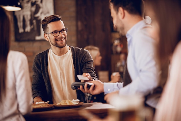 Szczęśliwy mężczyzna korzystający ze smartfona podczas dokonywania płatności zbliżeniowych w pubie