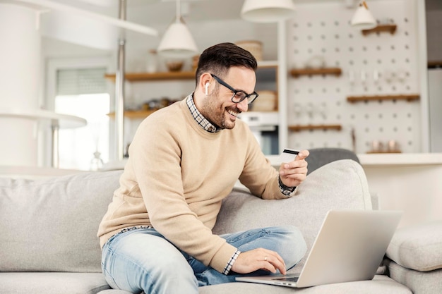 Szczęśliwy mężczyzna siedzący w swoim przytulnym domu i używający karty kredytowej i laptopa do zakupów online