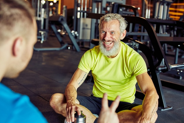 Szczęśliwy mężczyzna w średnim wieku rozmawiający o czymś z instruktorem fitness lub osobistym trenerem i uśmiechnięty