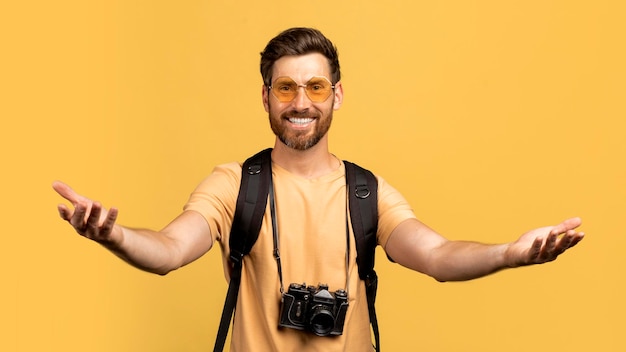 Szczęśliwy mężczyzna w średnim wieku w okularach przeciwsłonecznych wyciąga ręce do kamery stojący z żółtym plecakiem