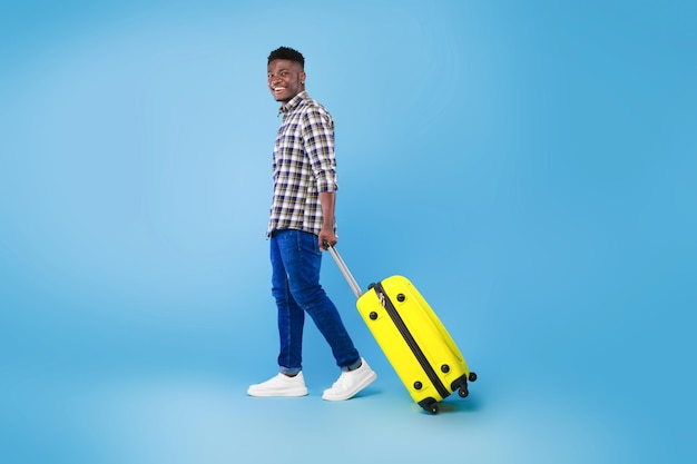Szczęśliwy młody afroamerykanin chodzący z jasną walizką na letnie wakacje nad niebieskim
