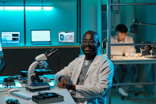 Szczęśliwy młody afrykański naukowiec z mikroskopem siedzący przy miejscu pracy