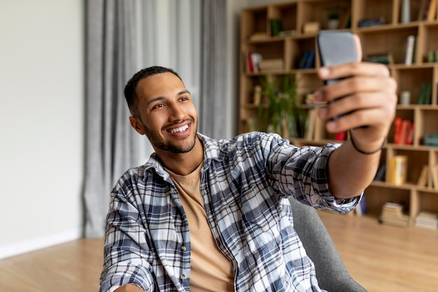 Szczęśliwy młody arabski facet rozmawiający przez smartfona lub robiący selfie siedzący w fotelu w domu