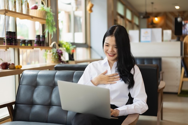 Szczęśliwy Młody Asian Kobieta Uśmiech Za Pomocą Laptopa Siedząc W Kawiarni. Młoda Kobieta Azjatyckich Siedzi W Kawiarni I Wideokonferencji Na Laptopie.