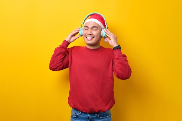 Szczęśliwy młody Azjata słuchający muzyki w kapeluszu Mikołaja ze słuchawkami stojącymi z zamkniętymi oczami na żółtym tle studyjnym celebracja świąt Bożego Narodzenia i koncepcja nowego roku