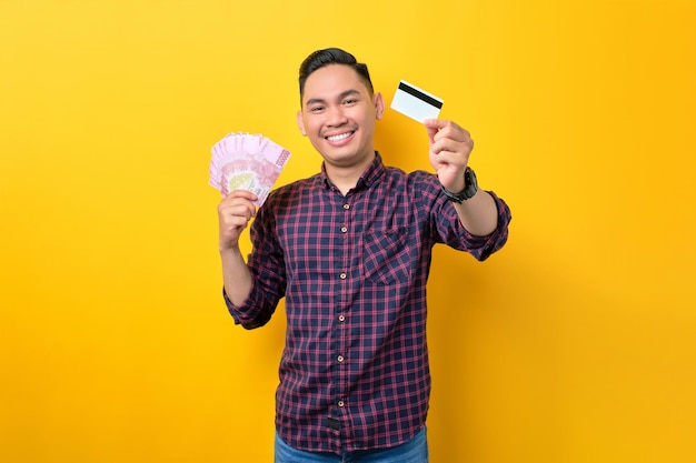 Szczęśliwy młody Azjata trzymający banknoty i kartę kredytową izolowane na żółtym tle Koncepcja łatwej płatności