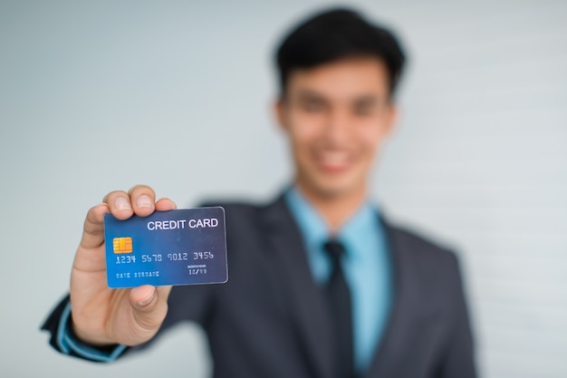 Szczęśliwy młody azjatycki mężczyzna przedsiębiorca uśmiechający się do kamery i demonstrujący kartę kredytową przed szarą ścianą