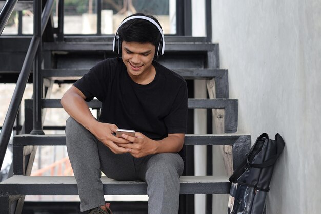 Szczęśliwy młody azjatycki student nosi bezprzewodowe słuchawki i uśmiecha się patrząc na telefon komórkowy