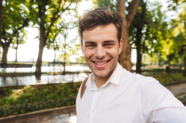 Szczęśliwy młody biznesmen na sobie koszulę, biorąc selfie, stojąc na świeżym powietrzu w parku miejskim