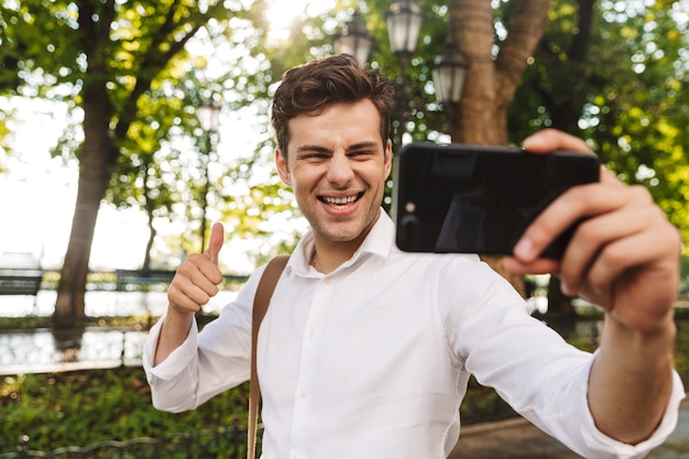 Szczęśliwy młody biznesmen w koszuli, biorąc selfie, stojąc na świeżym powietrzu w parku miejskim, trzymając telefon komórkowy z wyciągniętą ręką