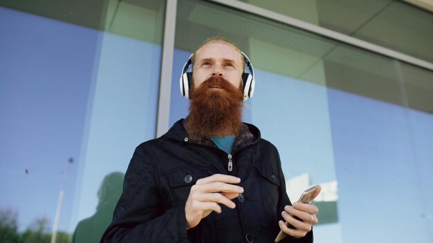 Szczęśliwy młody brodaty mężczyzna hipster ze słuchawkami i smartfonem, słucha muzyki