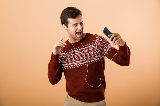 Szczęśliwy młody człowiek na białym tle nad beżową ścianą przy użyciu telefonu, słuchanie muzyki przez słuchawki i śpiew